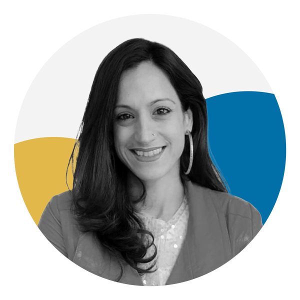 Elpida Kokkota, a woman with a light skin tone, smiles at the camera. Her portrait in grayscale. She has long black hair and is wearing a yellow jacket, white shirt, and coral lipstick. The background is a blue and yellow half circle.