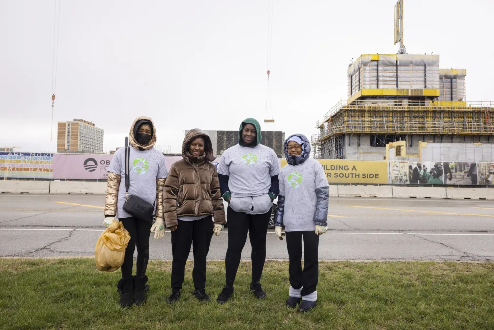 Under a cloudy sky, four Black teens with a range of light to deep skin tones stand in the grass in front of the Obama Presidential Center construction site in Jackson Park.