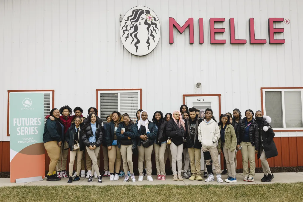 A group of students with medium-light to medium-dark skin tones, wearing khaki pants and a variety of hair styles, stand in front of a white building that reads “Mielle” and a graphic of a woman with long black hair on the wall.