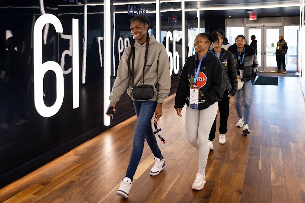 Young women with a range of medium to dark skin tones walk in a lit hallway. All are smiling. 