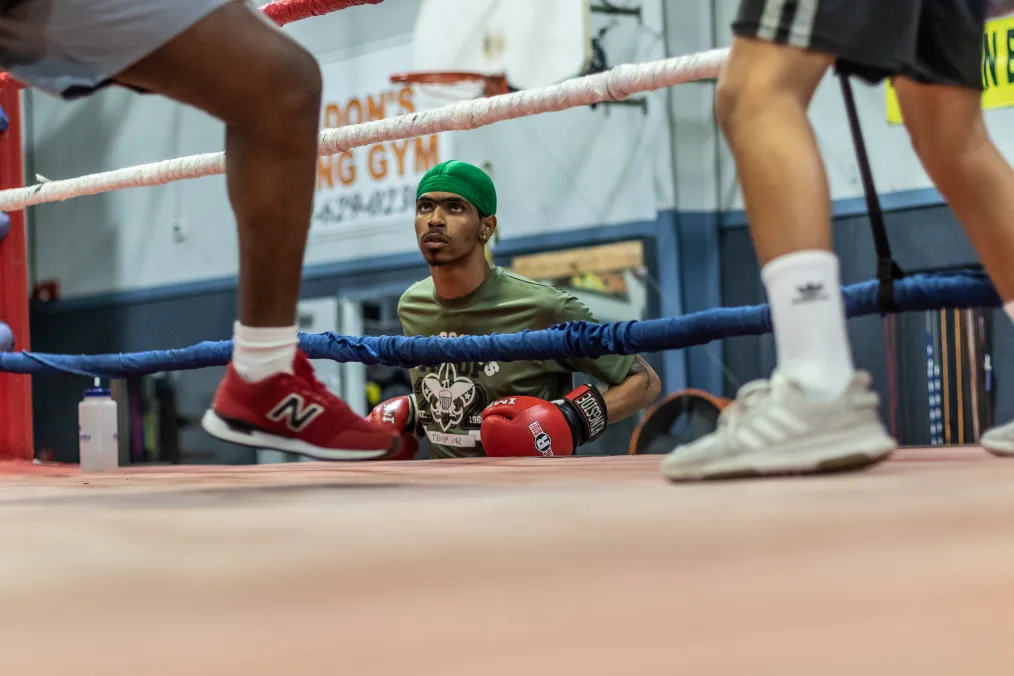 A man with medium-deep tone skin, a light green durag and red boxing gloves, looks into a ring with red white and blue boarders. In the photo frame are a pair of light-medium toned legs in fighting stance and a deep toned leg also in a fighting stance. Both have casual athletic attire on. 