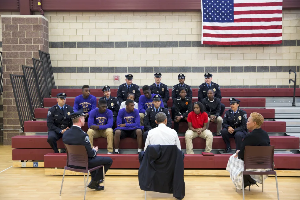 In a gymnasium, President Obama sits with youth and police officers for a town hall to discuss mental health and wellness. All have a range of light to deep skin tones. The officers are dressed in their uniforms.