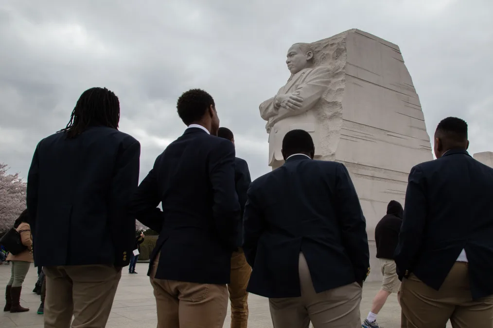 Four Black young men, dressed in dark suit jackets and brown khaki pants, are turned away from the camera, facing the Dr King monument in Washington, DC. The sky in the background is cloudy. 