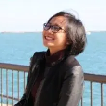 A light skin toned woman with shoulder length dark hair is smiling widely turned sideways to the left of the photo. She is wearing a dark red turtleneck sweater with a black jacket and black framed glasses. She is standing infront of a railing near a body of water. 