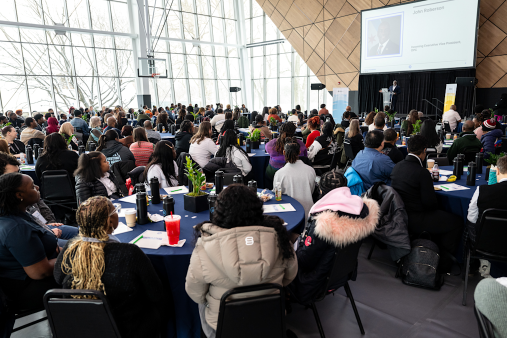 An audience sits at round blue tables in a large event room with tall windows. They watch a speaker standing on a stage next to a large presentation screen. The speaker is a Black man with a dark skin tone wearing a dark suit. The screen shows a photo and title for the speaker, John Roberson.