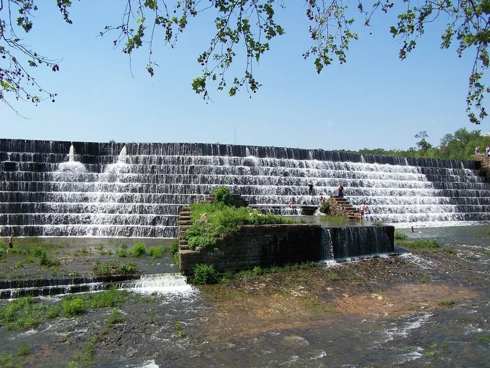 A large, tiered water feature with water running down it and green vegetation. There appear to be people walking on it in the distance.