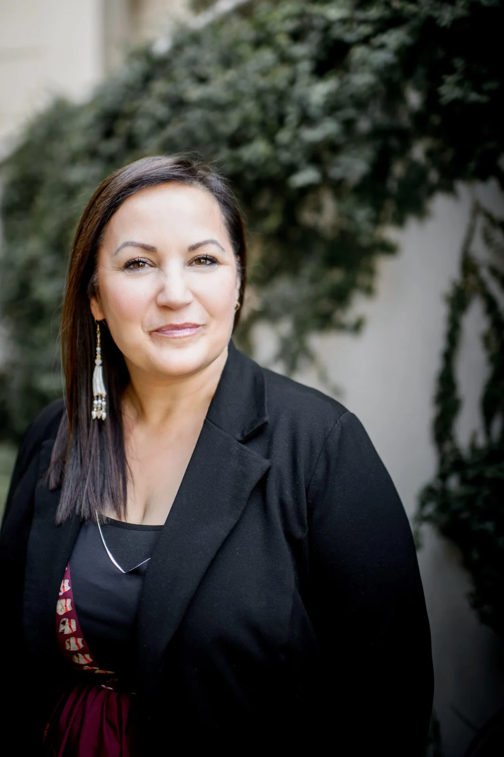 The headshot of a woman with light skin and straight brown hair, parted to the side. She wears a black suit jacket and dangling beaded earrings. Behind her is some green foliage.