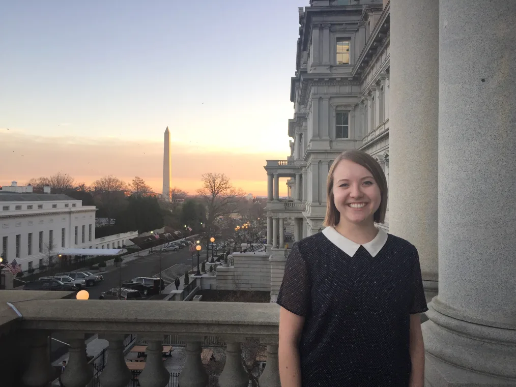 A photo of Kolbie Blume posing on a balcony with the Washington Monument in the distance. The sun is setting in the picture and the streets of Capitol Hill, Washington DC are in the background. Kolbie is standing on the balcony of a stone building with several windows. Kolbie has a pale complexion and light hair that falls to her ears. They are wearing a black shirt, with silver sparkles along it, and a white collar.