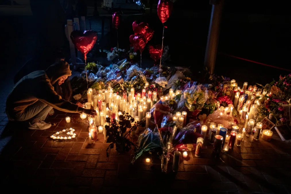 A person wearing a hoodie with the hood up is shown in profile, crouching down to light a candle. She is seen in profile in the glow of candlelight coming from dozens of votives surrounded by assorted flowers. A set of small votive candles are arranged in the shape of a heart near the person’s foot. Six heart-shaped red balloons float above the vigil. 