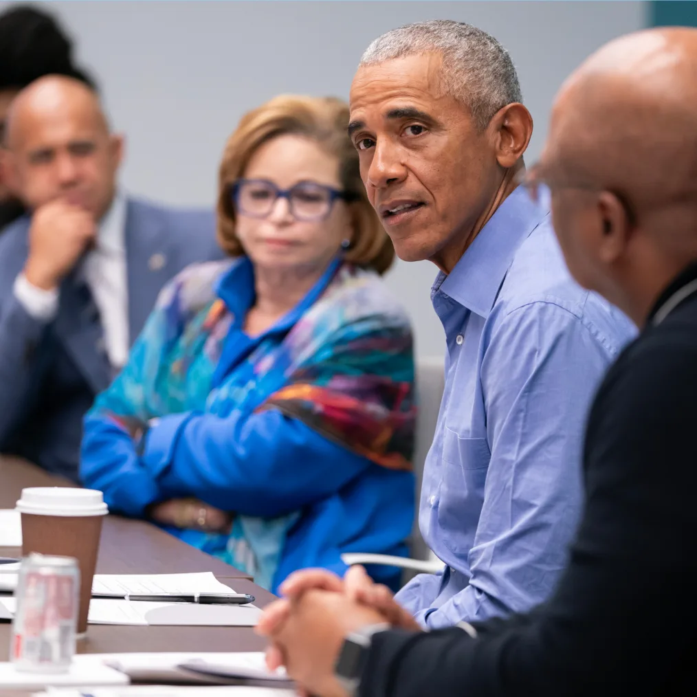 President Obama sits at a conference table addressing a group of people. President Obama has a light deep skin tone and gray hair. He is wearing a light blue button down dress shirt. Sitting next to him is Valerie Jarrett. She has a deep olive skin tone, light brown hair cut into a bob, glasses, and is wearing a multicolored blue jacket. 