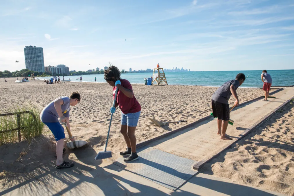 A group of people with various skin tones cleaning a beach with brooms and shovels.