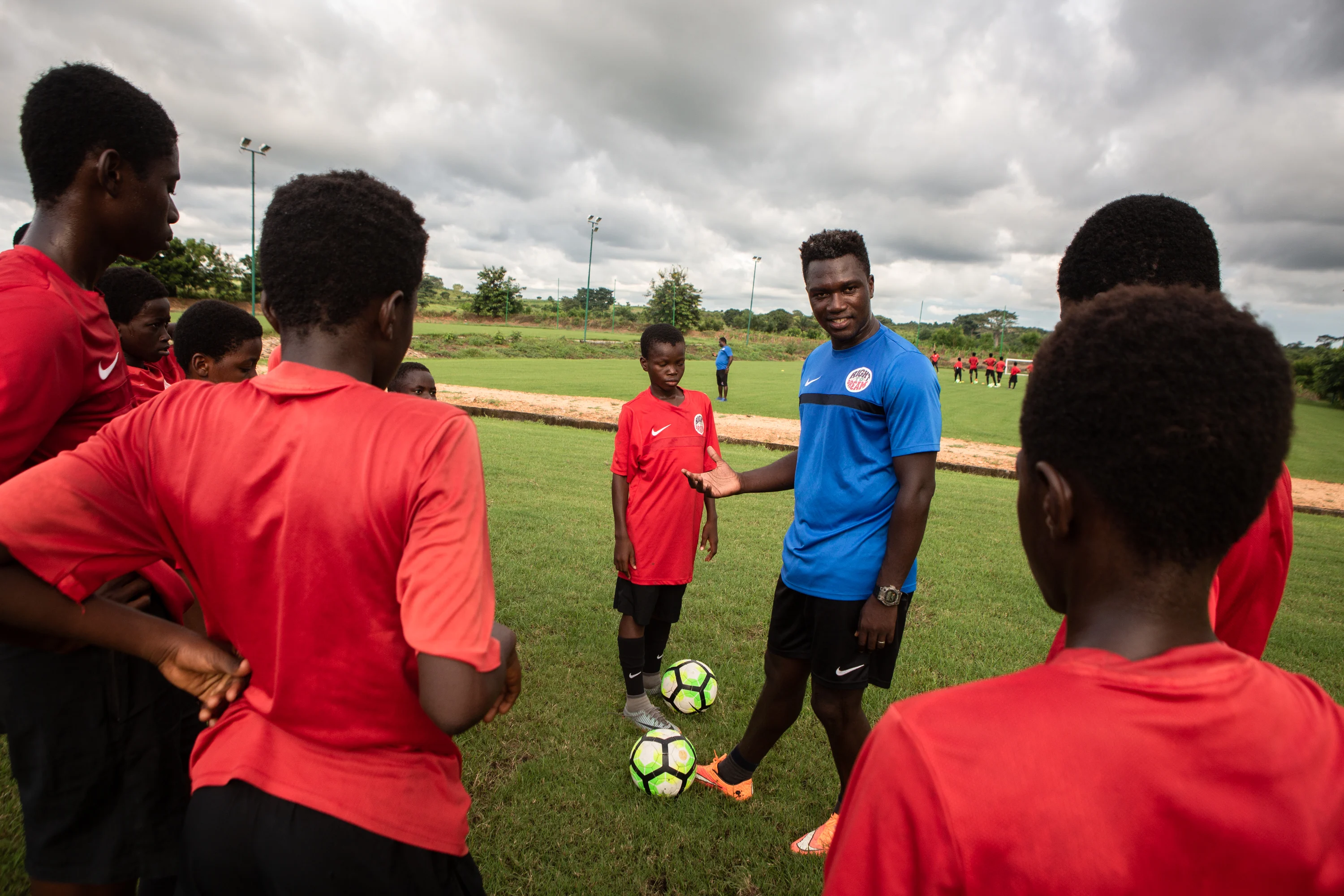 A team of  children with dark skin tones stand in a group around a coach teaching them how to play soccer 