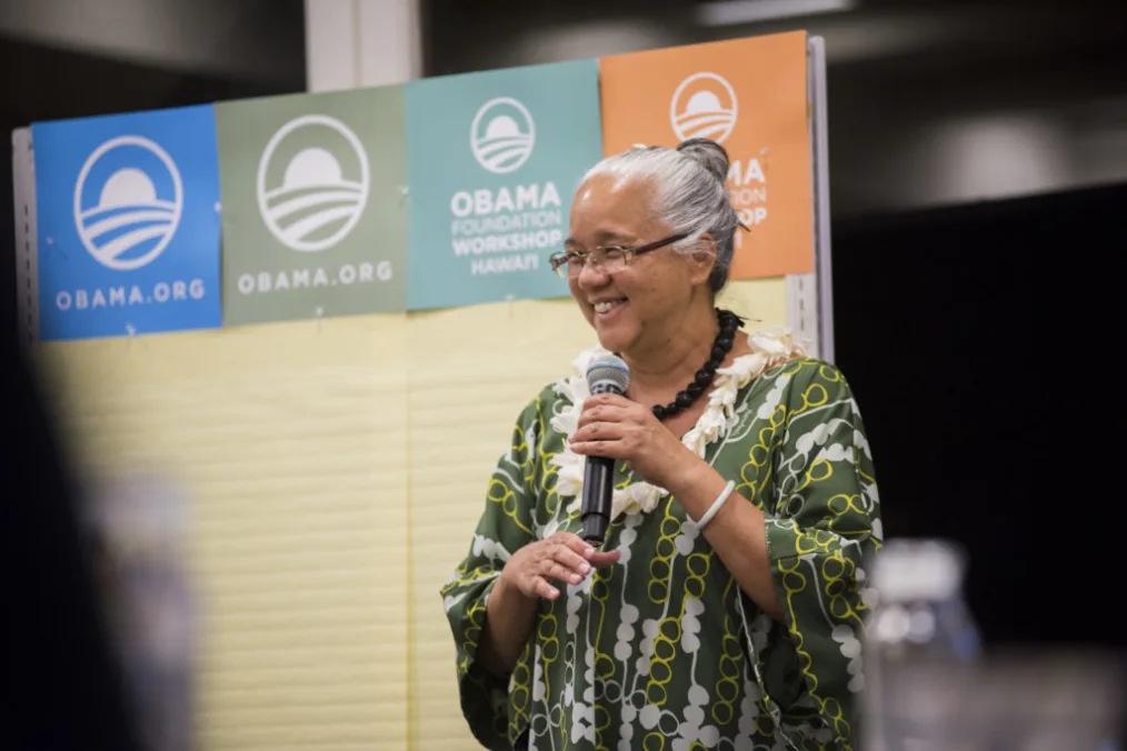 This picture shows a woman with a medium skin tone, short gray hair, and a microphone that seems to
be making a presentation on stage with Obama Foundation logos in the background.