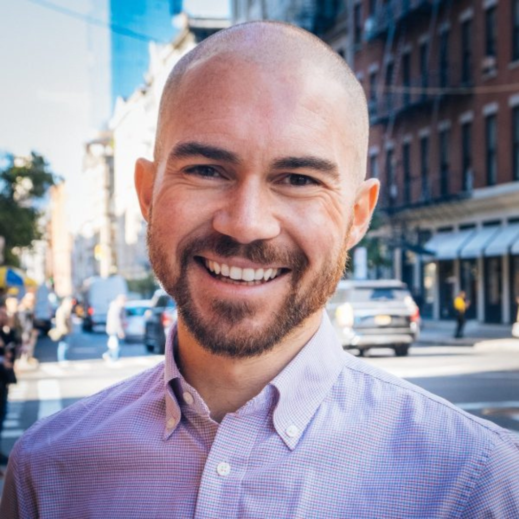 The image is a headshot of Jason Rahlan. He has a pale complexion, dark eyes, a bald head, and a dark brown goatee. He is wearing a checkered shirt and smiling into the camera. Behind him is a city street (presumably a street in Manhattan, New York.)