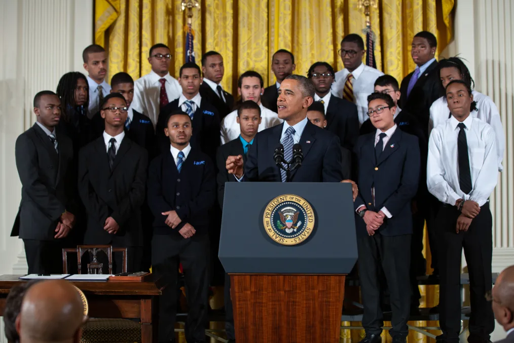 President Obama speaks from behind a podium at the White House. Behind him are a group of 20 young men. They are a range of light to deep skin tones and are dressed professionally. 