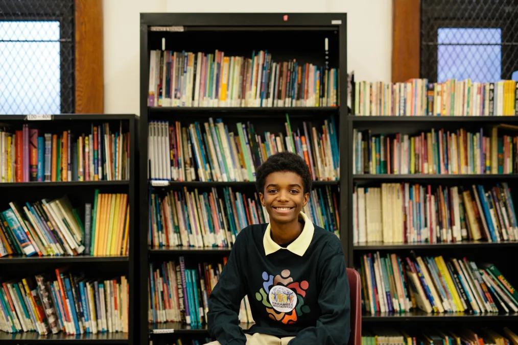 A young Black man smiles at the camera as he sits in a chair in front of bookshelves filled with books. He wears a sweatshirt with the words "Harambee Brotherhood."