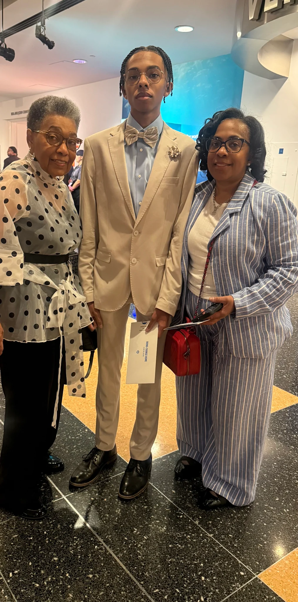 Jonathan Sullen, a 16-year-old Black boy with a light skin tone, stands in between his mother and grandmother. Both are Black women with medium skin tones. All are dressed professionally. 