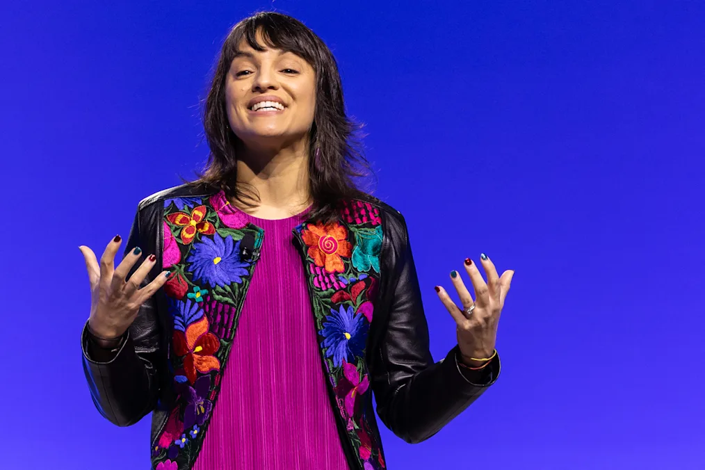 Mónica Guzmán, a woman with a light skin tone smiles as she speaks to an audience at the 2024 Democracy Forum. She has short brown hair and bangs. She is wearing a pink top and multicolored leather jacket.