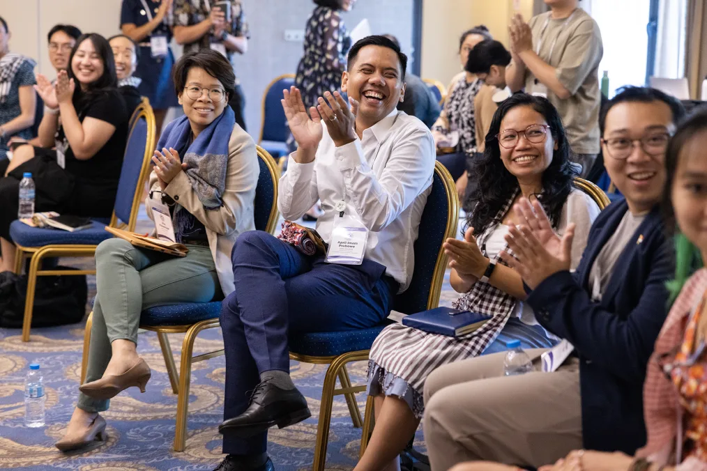 A group of Obama Asia Pacific leaders smile and clap as they sit on chairs in a conference room. They are all dressed professionally. 