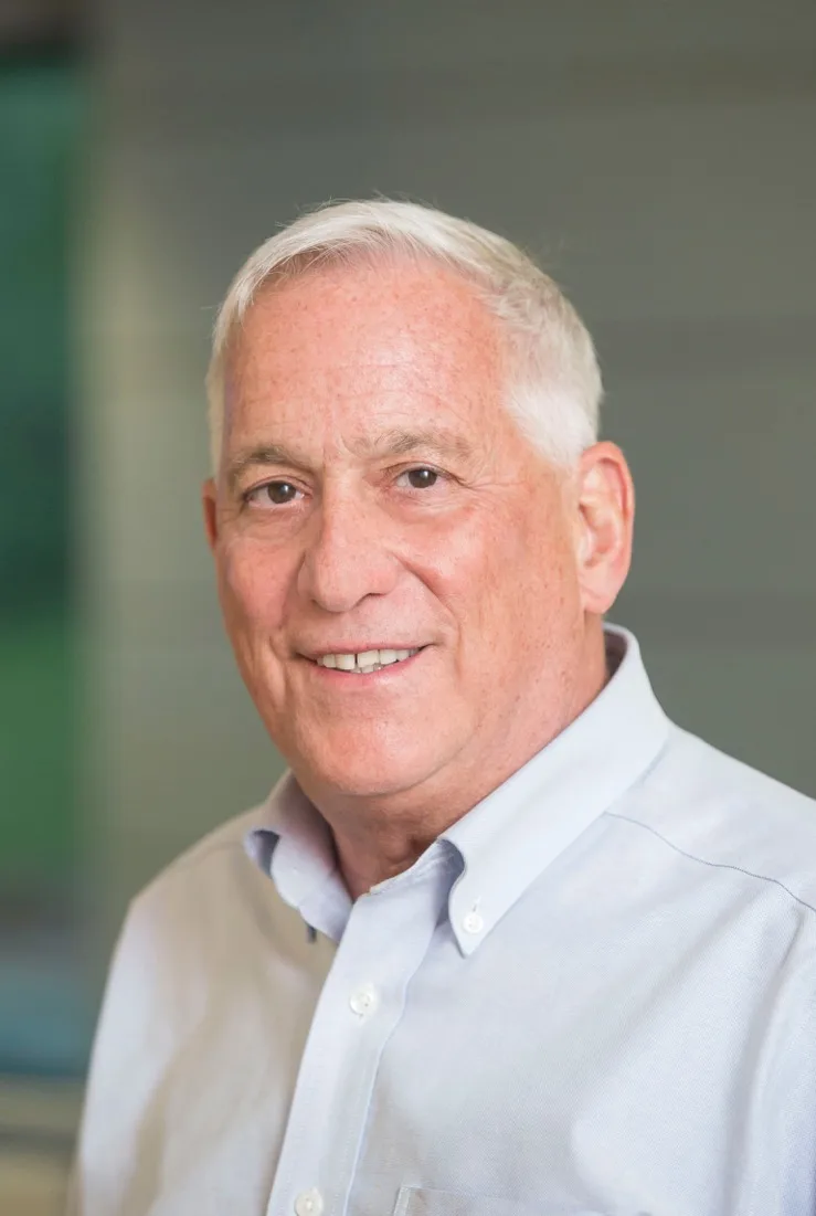 A headshot of an older White man with white hair smiling slightly for the camera. He wears a light blue button-down shirt.