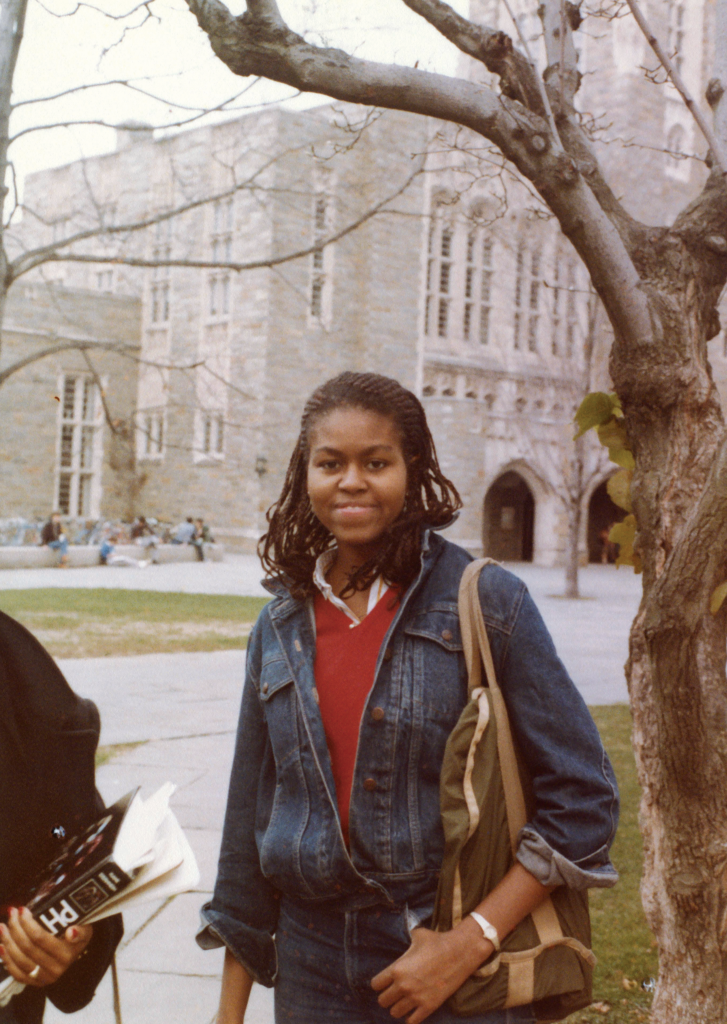 A young Michelle Obama stands on the campus of Princeton University. She smiles with long and wavy braids, is wearing a jean jacket anda bright red shirt. 