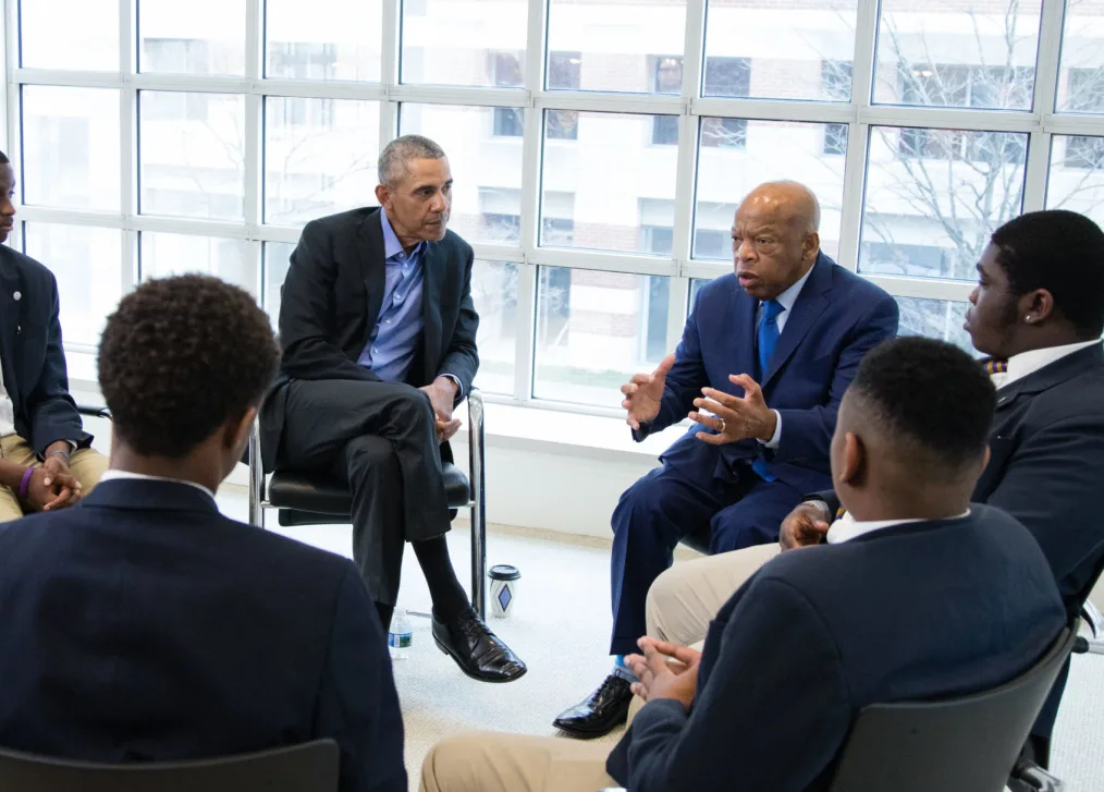 Barack Obama sitting and looking at a man with a light medium brown skin tone talk to other boys with brown skin tone in formal suits