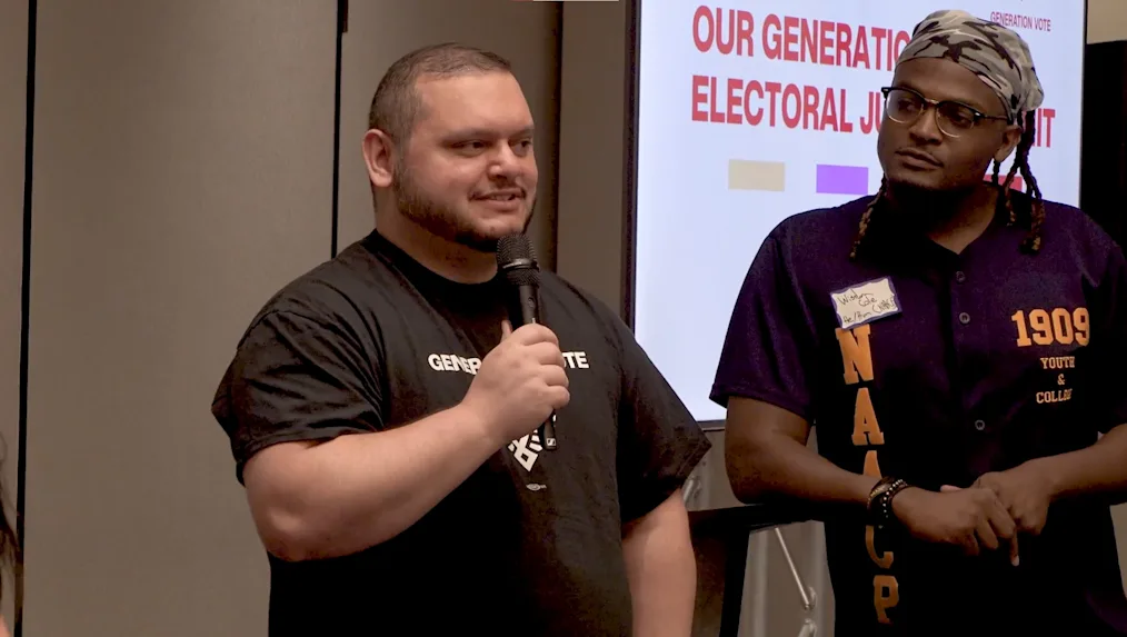 Murat Akaydin, a Turkish-American man with a medium skin tone, holds a microphone as he speaks. A Black man with a dark skin tone stands beside him.
