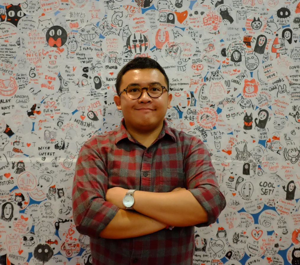 A man with a medium skin tone has a short haircut, glasses, and is wearing a red and gray plaid shirt. He smiles as he stands in front of a whiteboard with various abstract designs in black, blue, and orange colors.