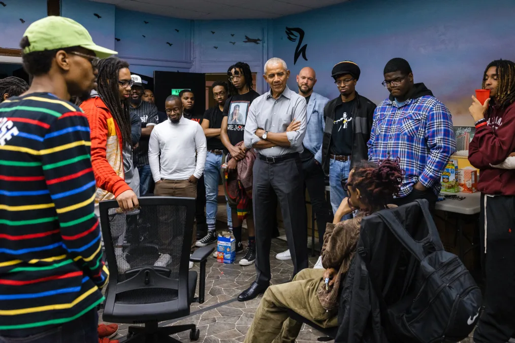 President Obama stands in the middle of a recording studio. He is surrounded by 15 other young people with a range of light to deep skin tones. President Obama has his arms crossed and is wearing black slacks and a gray button down shirt.
