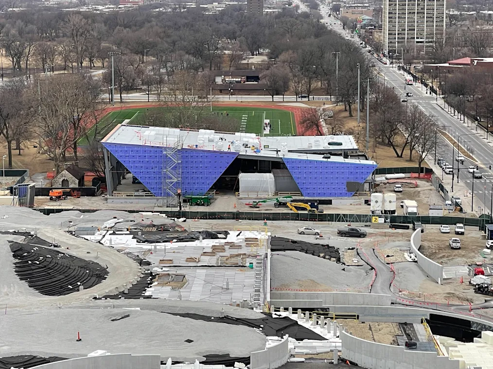 Two bluish purple inverted triangles of wall substructure look to be wedged into the ground on the north face of Home Court, hinting at the shape of the metal panels that will eventually cover this part of the building, and the shape of glass walls between the wedges. In the foreground concrete angular and curved support walls and railings, and contours of geofoam span the landscape where the playground and part of the great lawn will eventually be.