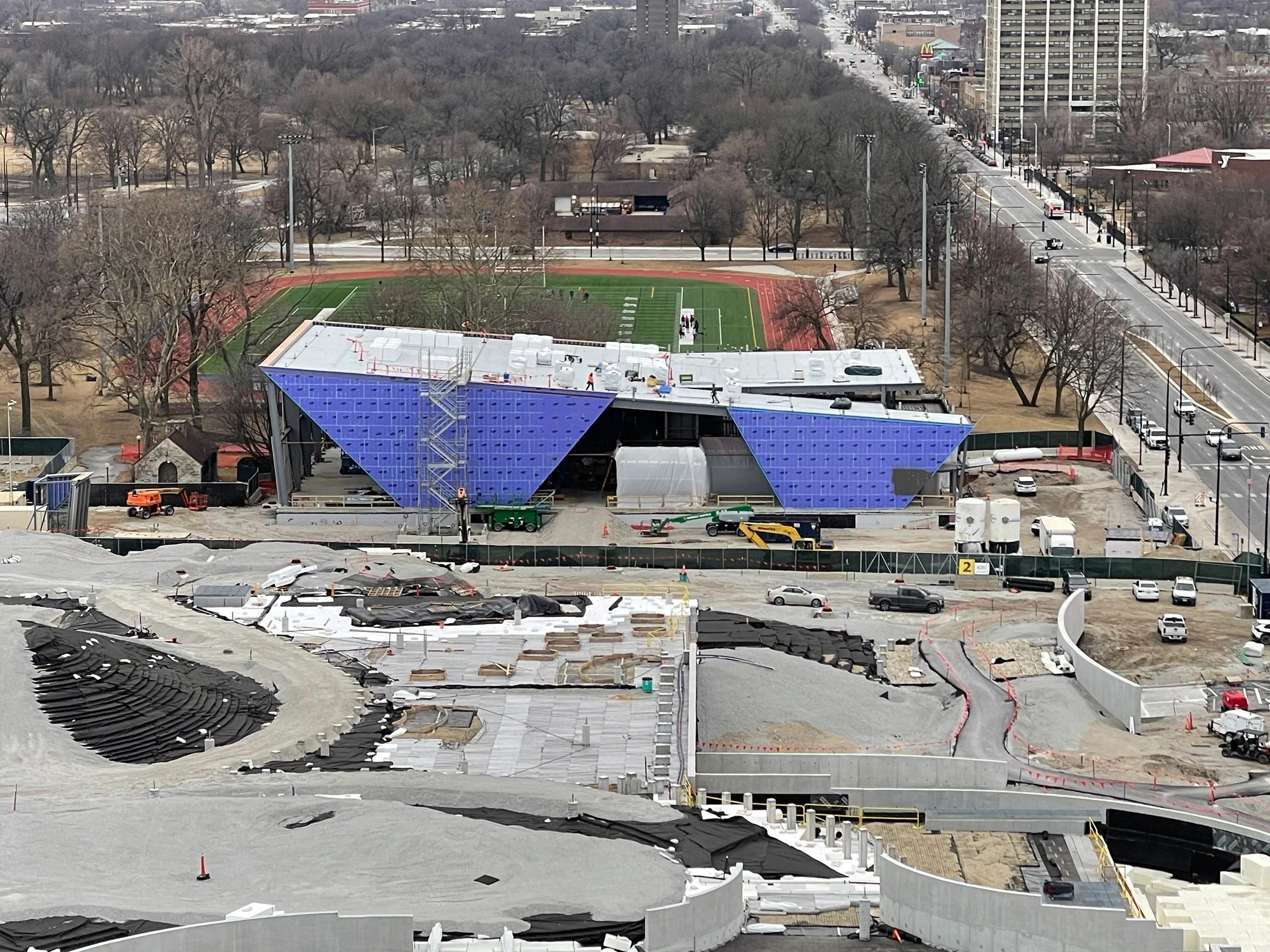 Two bluish purple inverted triangles of wall substructure look to be wedged into the ground on the north face of Home Court, hinting at the shape of the metal panels that will eventually cover this part of the building, and the shape of glass walls between the wedges. In the foreground concrete angular and curved support walls and railings, and contours of geofoam span the landscape where the playground and part of the great lawn will eventually be.