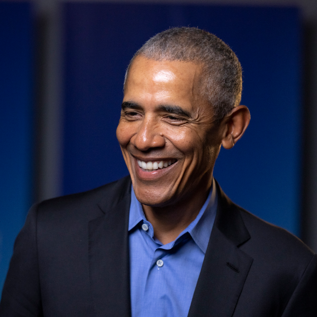 President Obama smiles in front of a blurred blue background. He is wearing a suit jacket and blue shirt.