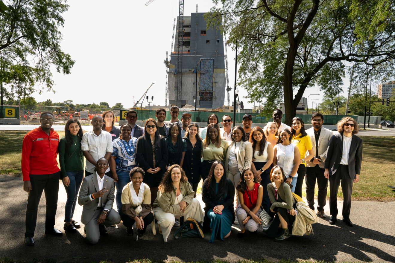 A group of adults, varying in skin color and age stand and kneel in front of the Obama Presidential Center as it is under construction. Cranes and scaffolding can be seen around the building.