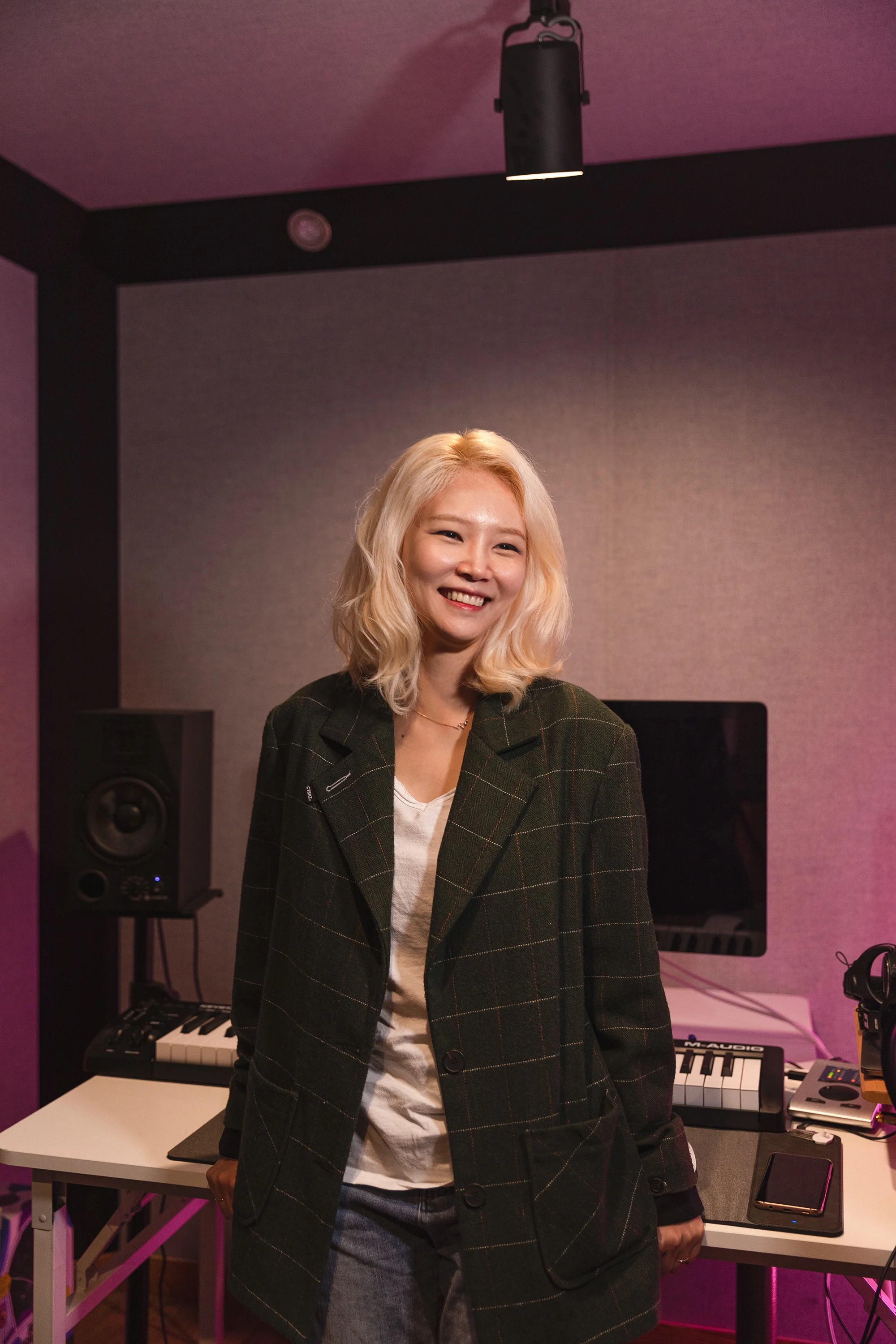 Michelle Cho, an Asian woman with a light skin tone and platinum blonde hair, smiles as she stands in front of a keyboard in a studio.