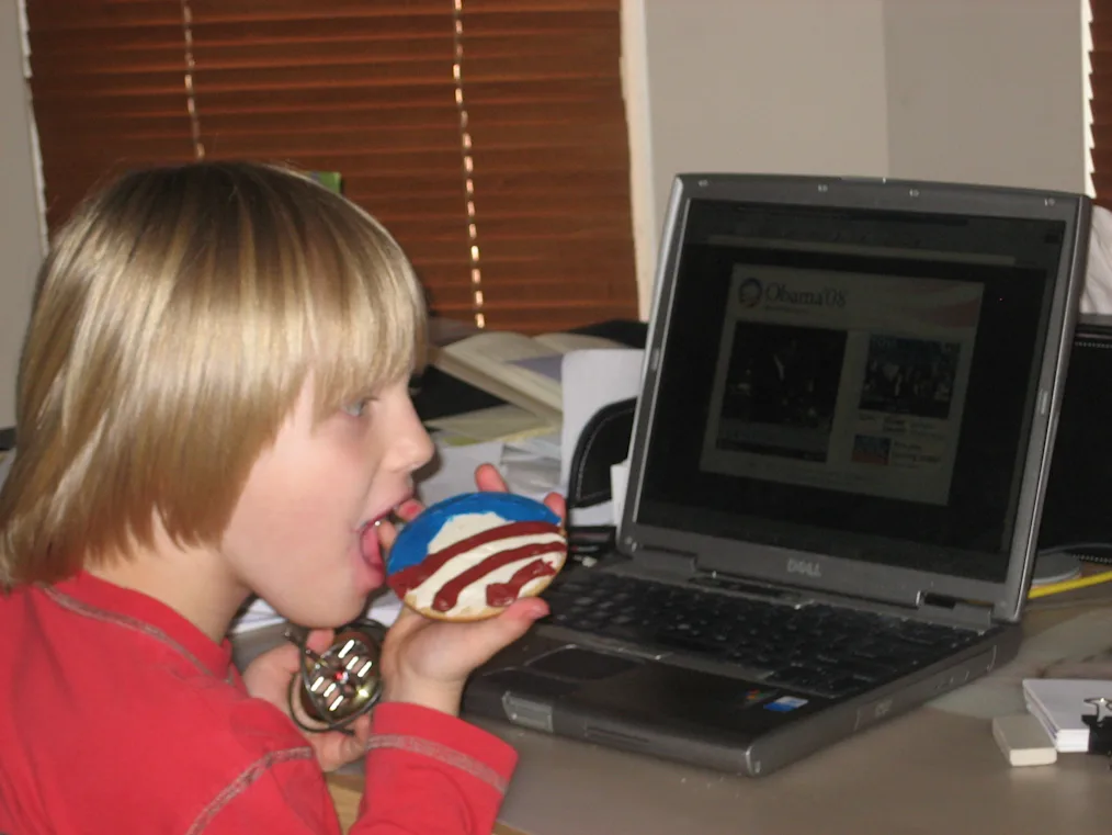 A young boy with blonde hair and bangs clicks on a cookie with the Obama logo. He sits at a table with a laptop in front of him. 