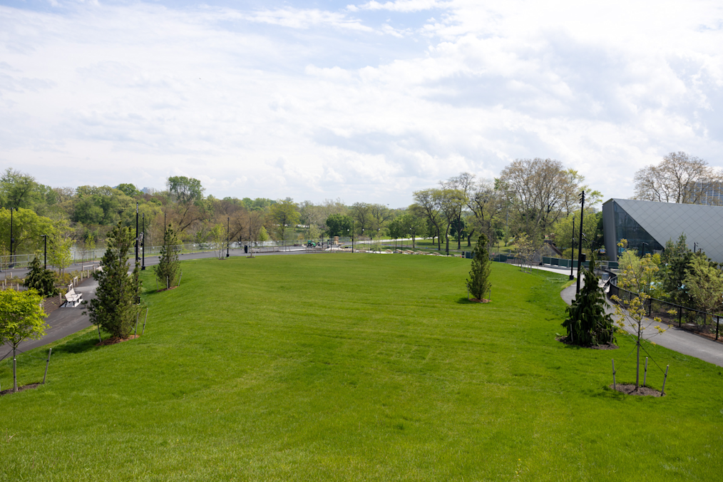 A wide green lawn slopes gently downward toward a paved walking path surrounded by trees under a cloudy sky. On the right, a modern silver building sits beyond a small paved walkway.