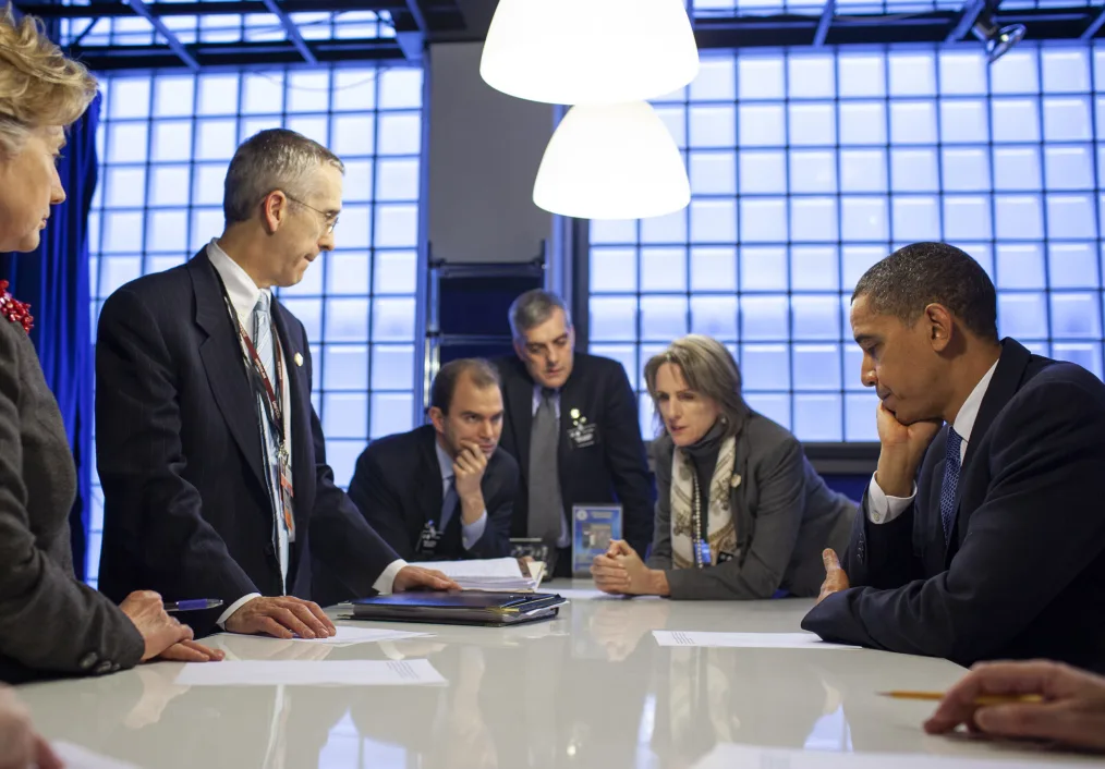 President Obama sits at a long white counter looking over a document with other older men and women with light skin tones. They are sitting around him in business attire  