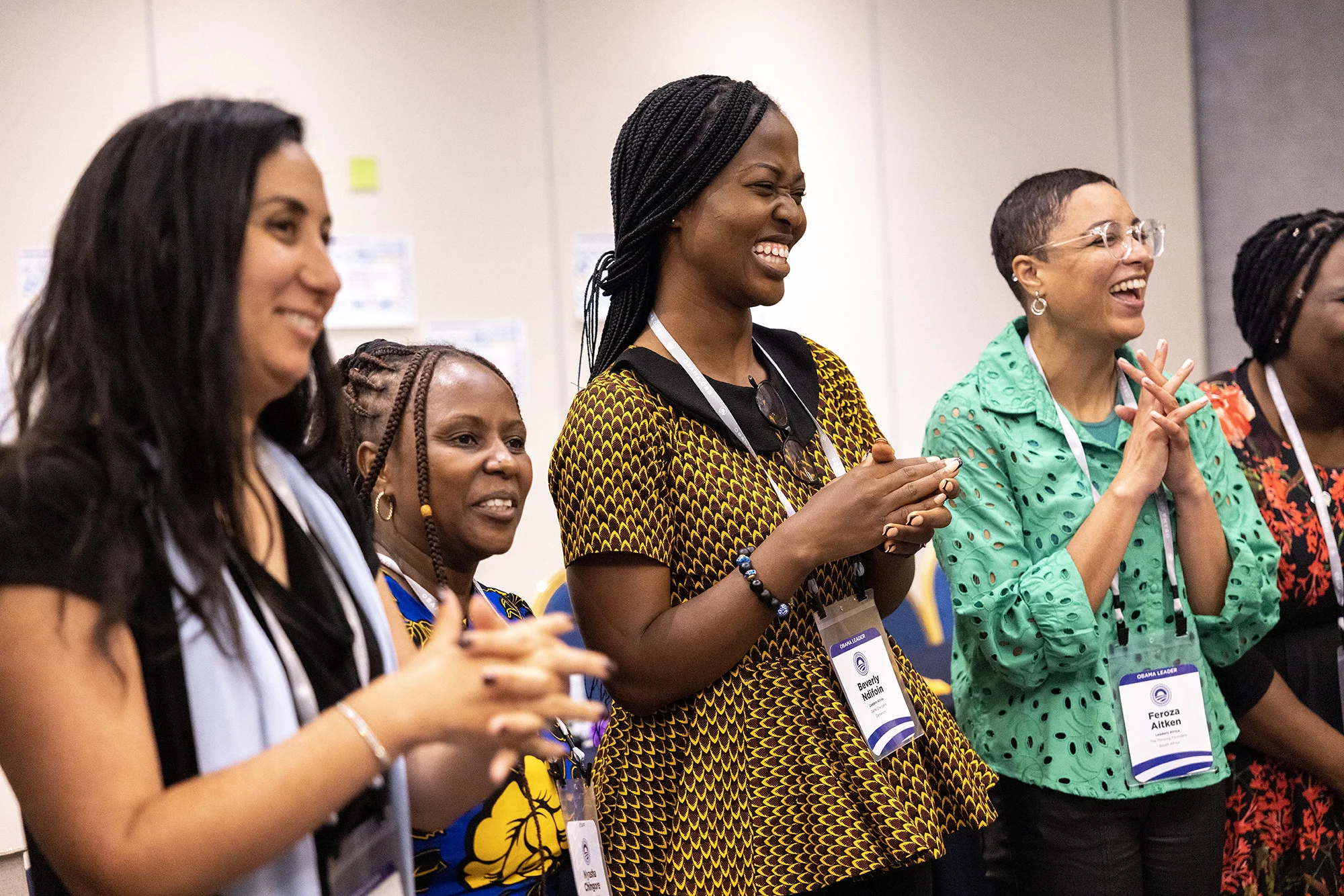 Five women with a range of light to dark skin tones smile and clap as they look away from the camera. 