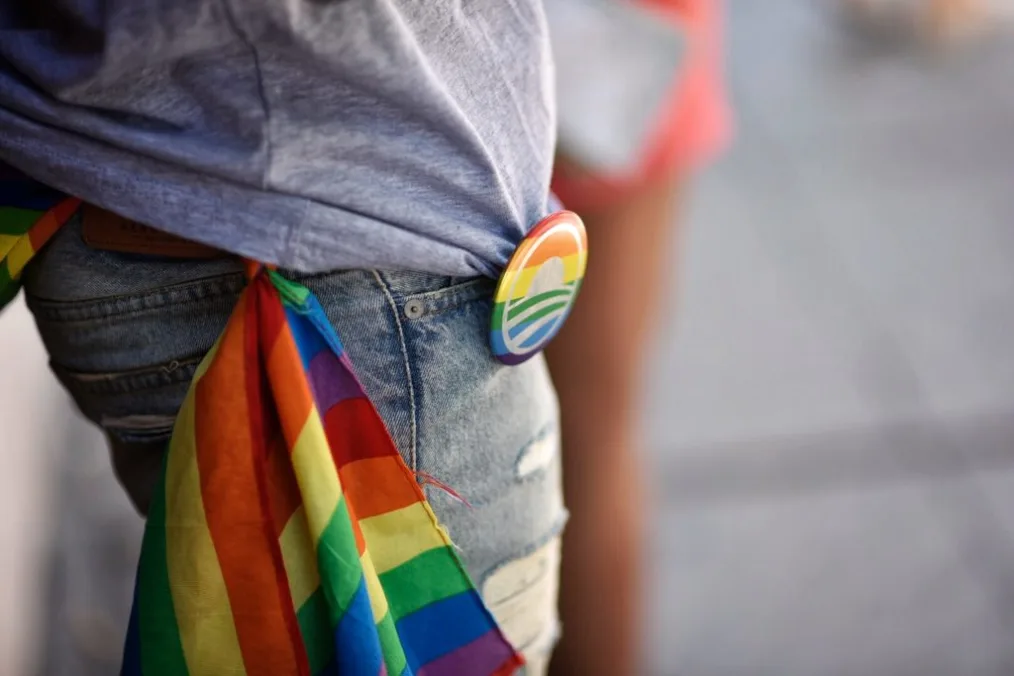 
A zoomed-in image of someone's jeans and a t-shirt with a pride flag hanging out of the back pocket and an Obama pride-colored logo pin attached to the shirt 