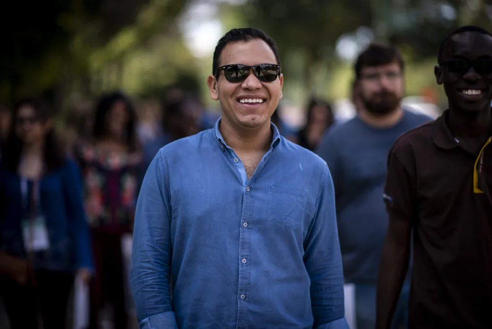 The camera focuses on a man with a light medium skin tone, black sunglasses, short black hair, and blue button up walking down a street as he smiles