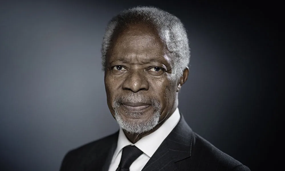 Kofi Annan, a black man with a deep skin tone, short grey hair, and grey beard wearing a black suit and white collard shirt, looks into the camera.