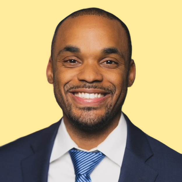 The image is a headshot of Derick Dailey. Derick has a medium deep skin tone, closely cropped black hair, brown eyes, and a dark brown goatee. Derick is smiling in the photo. Derick is wearing a dark blue suit jacket with a striped tie that is black, blue, and white. Underneath the suit jacket and tie is a white collared shirt. The background of Derick's image is the color yellow.