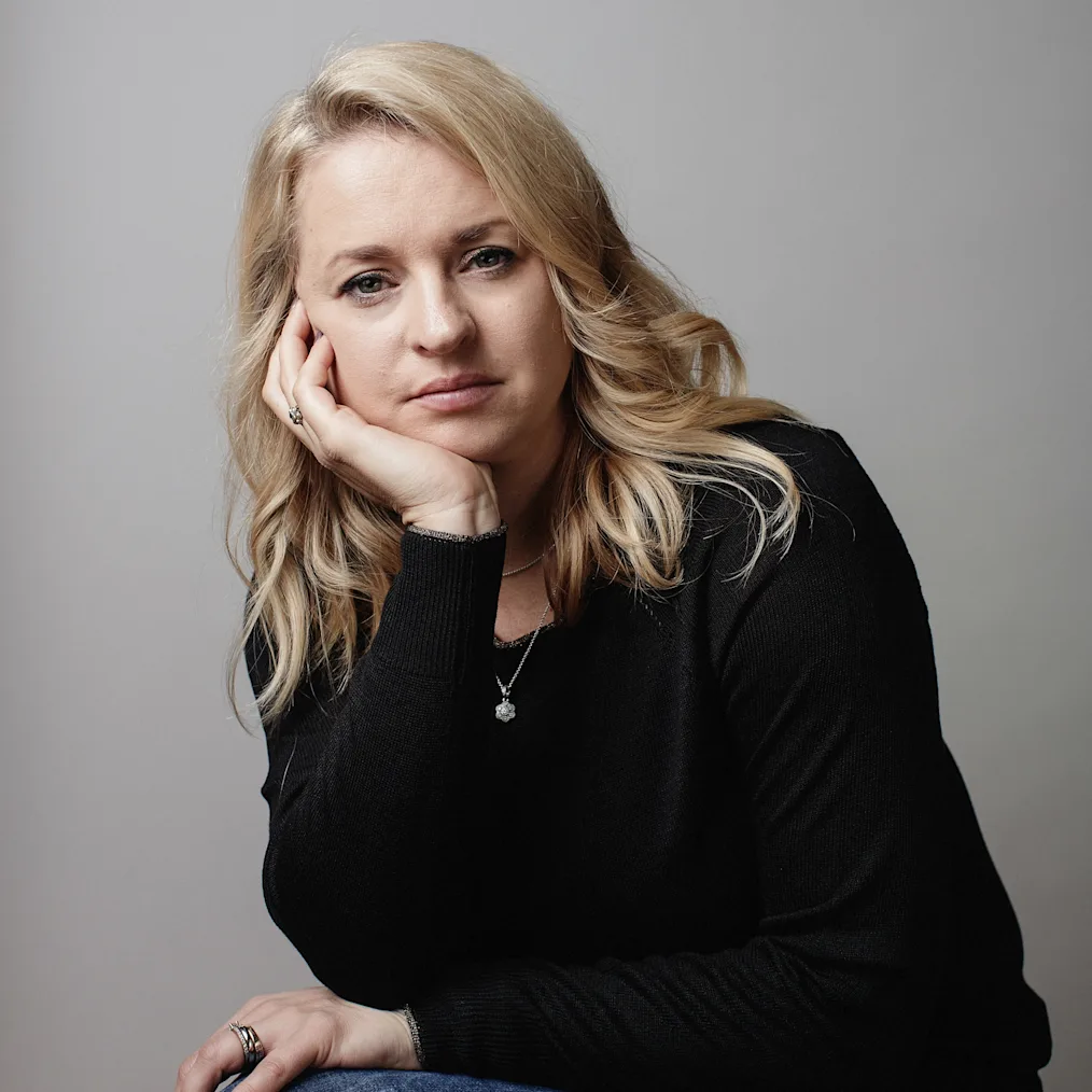 Tammy Darcy, a woman with a light skin tone, holds her face in her hands as she poses for a portrait. She has blonde hair and is wearing a black sweater and blue jeans. 