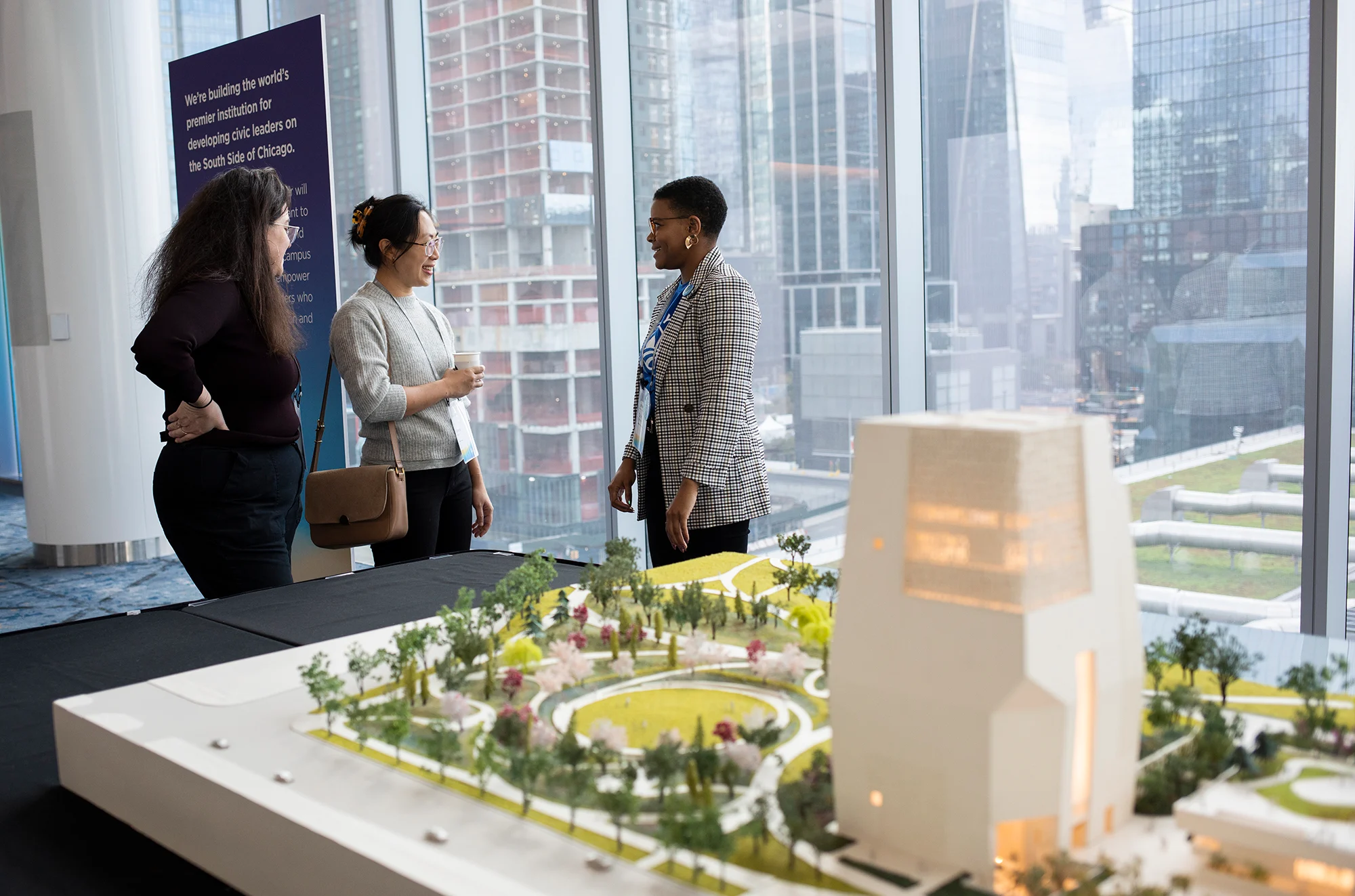 Three women, with a range of light to medium dark skin tones, stand and talk amongst each other next to a physical model of the Obama Presidential Center campus. All are dressed professionally. 