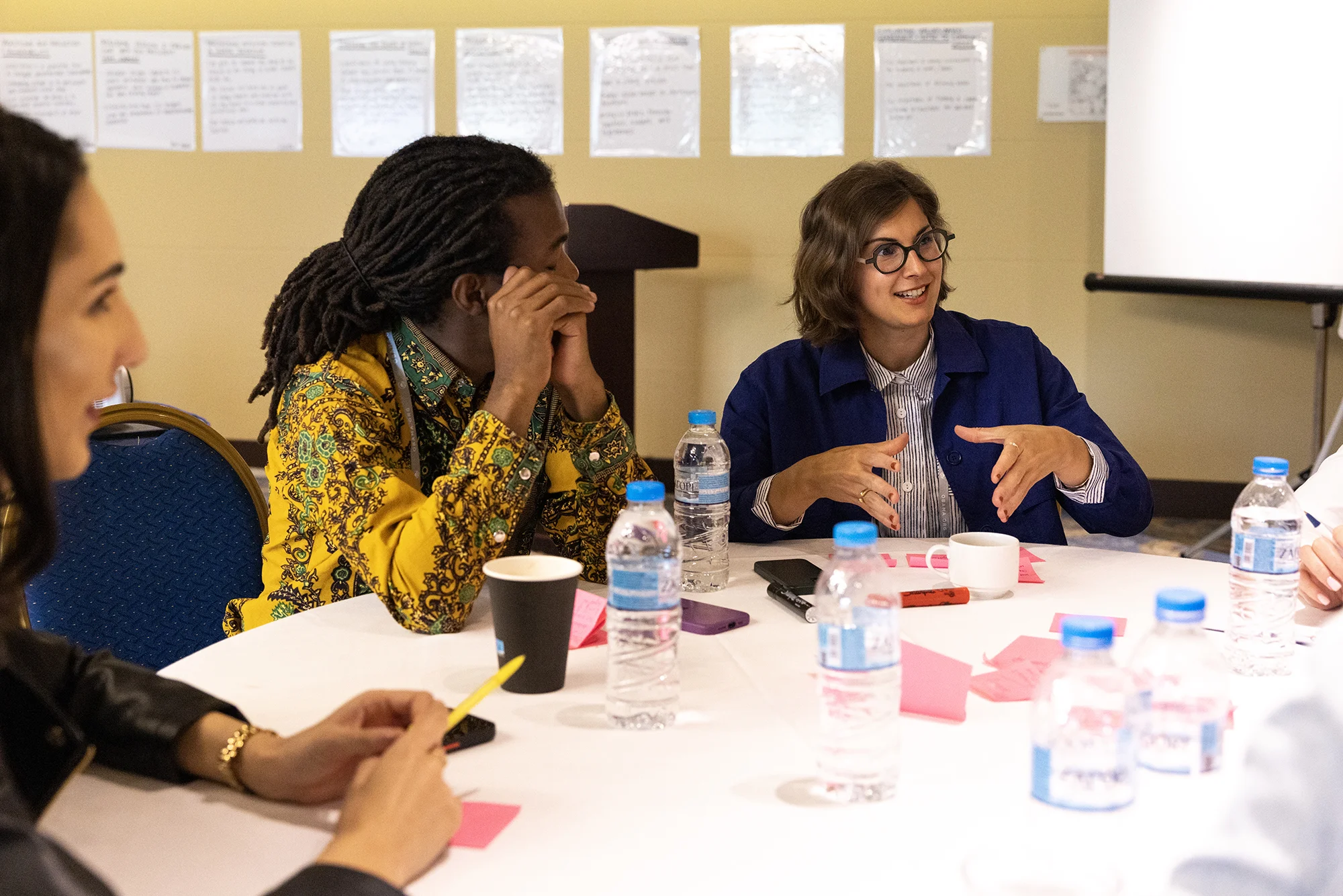 A man with a deep skin tone clasps his hands on the right side of his face looking at a woman with medium-light skin, as she gestures and smiles. They sit at a white table covered with sticky notes, bottles of water, and other materials.