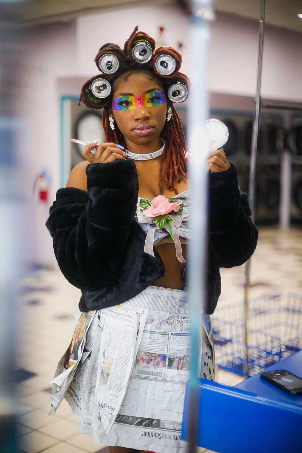 An image of a woman in a public laundromat. The women has a medium deep complexion and red locs and hang to her shoulders. The red locs at the top of her head are rolled in soda cans. Across her eyes is a painted pride flag. She also has dangling diamond earrings. She is wearing a black fur coat that is hanging off of her shoulders and a bra top and skirt made of newspaper clippings. In her hands she is holding a hand held mirror and a lip gloss wand. Behind her are a set of industrial washing machines. Next to her is a blue laundry folding station. On top of the blue station is a blue Iphone.