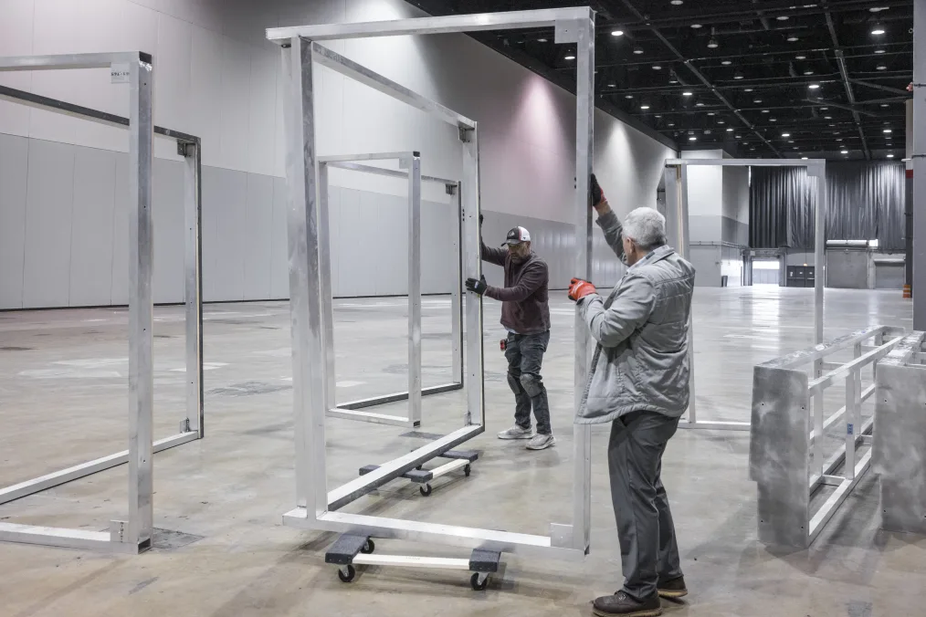 An image of the construction site of the Power of Words display at the Obama Presidential Center Museum. In the photo, two people with light skin tones are steadying a silver metal display beam on two rolling carts. The display beam is about seven feet tall. The person holding the left side of the beam has short gray hair and is wearing orange construction gloves, a silver jacket and gray pants. The person holding the right side of the beam is wearing black construction gloves, a baseball cap, a dark colored shirt, and gray pants. To the left of the two people is another beam sitting upright on the floor of the construction site. On the right side of the two people are silver beams laying down on the floor of the construction site. In the distance, behind the two people, is an open door next to a staging site covered by silver curtains. On the ceiling of the location are black steel rods with spotlighting.
