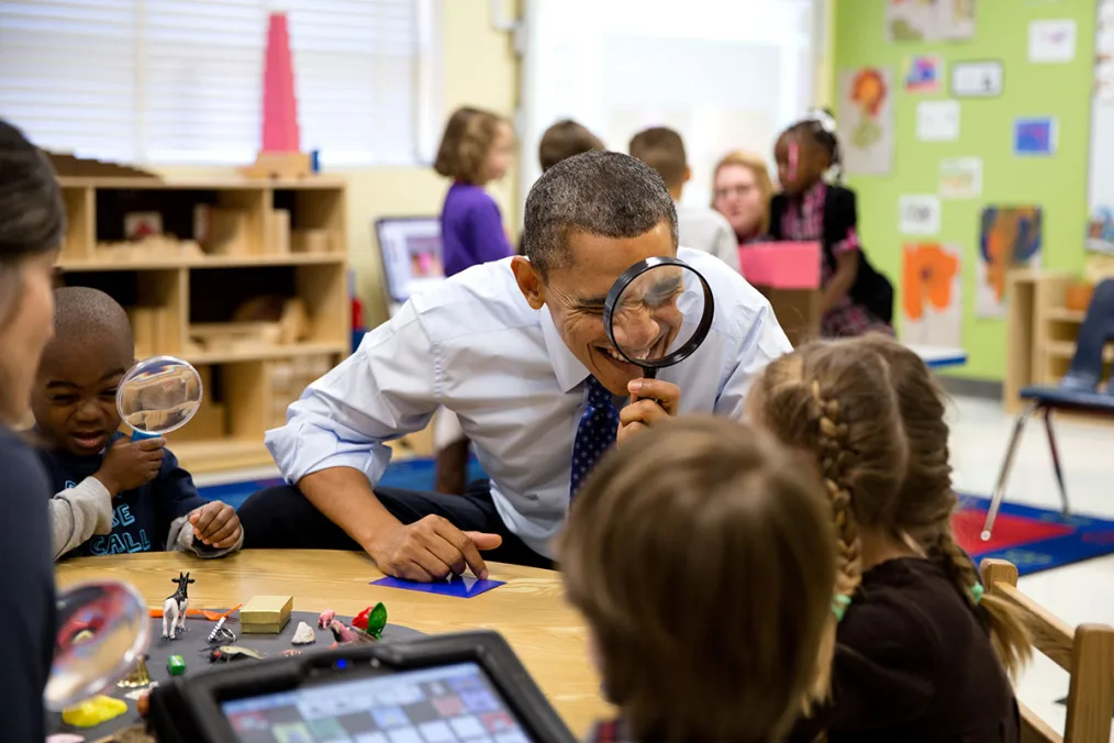 President Obama in a classroom with multiple children around him looking through a magnifying glass
