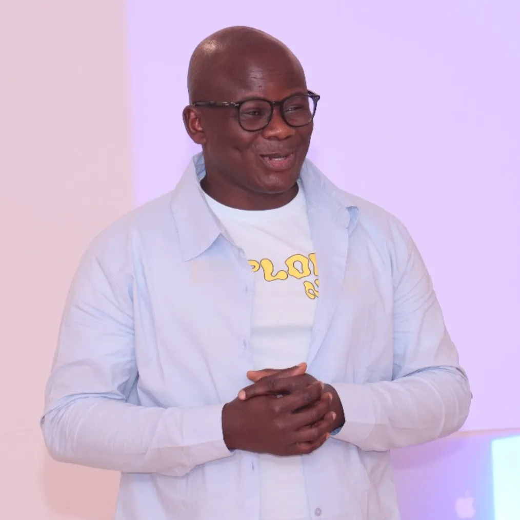 Paul Ekuru, a Black man with a dark skin tone and no hair, speaks to an audience. A sign in the background reads, “The Pawlehouse Company.”