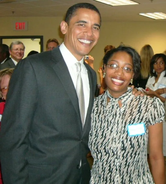 Barack Obama, wearing a suit and tie, poses for the camera with his arm on the shoulder of a Black woman wearing a patterned dress and name tag. They appear to be at a gathering, as people can be seen milling around behind them.